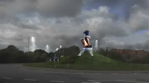 Peter W Naylor/ERYC Planning Portal An artist's impression of the sculpture on a plinth with the words "Three Bags Full" on the side. It is on a grass mound. The bronze bags have wool sticking out of them. The black metal sheep is standing on the top bag. It is night-time and is lit with floodlights from below.