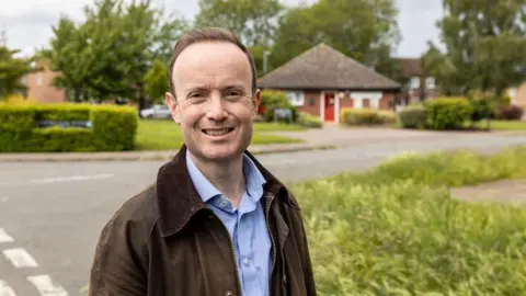 Jo Thewlis/BBC Richard Rout standing in front of hedges with a road and bungalow behind him