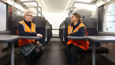 Two women wearing orange hi-vis vests are sitting at tables inside the HS2-mock carriage looking at each other across the aisle. 