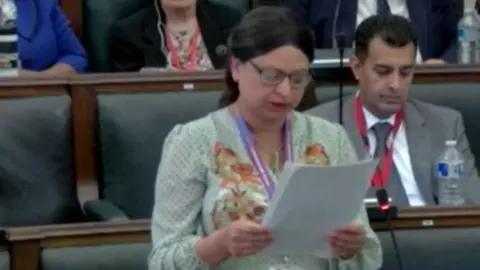 Birmingham City Council A woman standing in a council chamber, reading from a paper. She is wearing a white dress with green spots and floral embroidery, and has dark brown hair tied back in a centre parting, and glasses. Behind her are rows of green upholstered seats. A man sits in the row behind, in a grey suit, white shirt and grey tie, with a water bottle in front of him.
