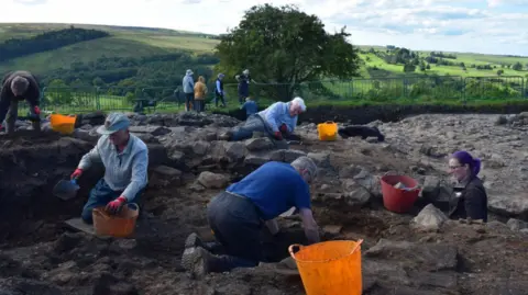 The Vindolanda Trust Lots of people in shirts and caps digging in the ground with orange buckets next to them. There are green hills and a large tree with greenery in the background.