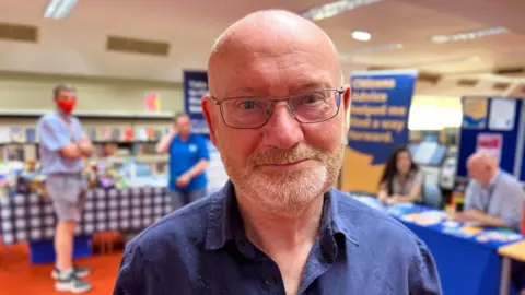 A man wearing glasses and a dark blue shirt. He is bald with a grey trimmed beard and is standing in a community centre with an orange carpet and blue tables with support stands set up.