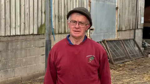 BBC Older man wearing glasses and a hat with a red jumper on, standing in a farmer's shed. Straw can be seen on the ground behind him and coop panels stacked against the wall.