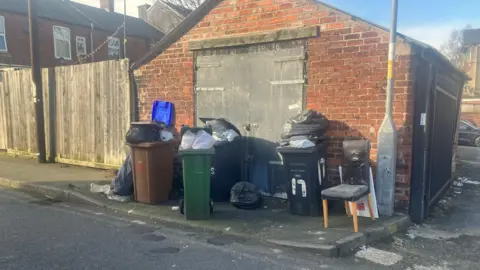 Photograph of overflowing bins and furniture which has been left on the street - outside a garage - in the Dukinfield area of Tameside.