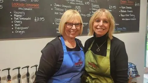 Handout Volunteers stand in the cafe under the menu board. Penny is wearing a blue apron and Julie is wearing a green apron. They are standing in front of a chalk  board with a menu on it.