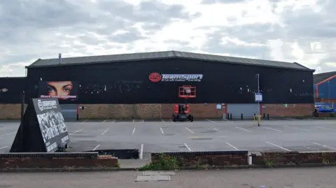 The building is black and brick with its sign on one wall in white writing. There is a car park in front of it.
