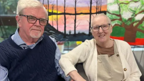 Sally and Kevin are looking at each other and smiling. Kevin is wearing a blue and white striped shirt and a navy knitted tank top. Sally is wearing a cream cardigan, brown t-shirt and light brown dungarees. Both have short white hair and glasses.