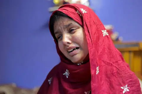 SYED SHAHRIYAR / BBC A woman wearing a red headscarf with white floral embroidery, standing against a blue background with blurred objects behind her.