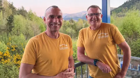 Lochaber MRT Iain Murray and Astie Cameron, wearing orange Lochaber MRT-branded T-shirts, are standing in the outdoors. There is a stunning view to trees and mountains behind them.