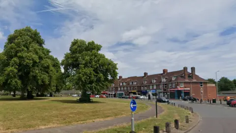 Picture shows a large green roundabout, with two large full green trees in the middle. To the back right are a row of shops in red brick terraces with homes above them. 