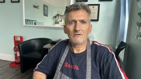 BBC/Melanie James Lee seated in a black salon-style chair indoors, wearing a navy T-shirt with red “Bench.” text and a grey apron. The room has pale blue walls, framed certificates, a wall mirror, a black armchair, and a red fire extinguisher visible in the background.