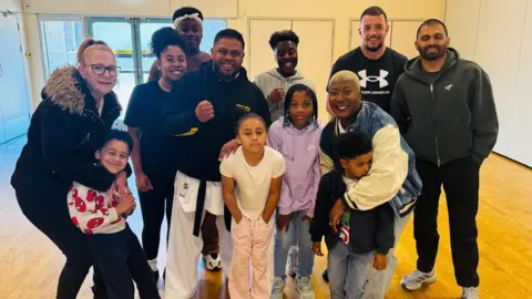 Shelim Ali A group of adults and children are posing for a picture inside of a hall. Some are wearing martial arts kit and are clenching their fists. Others are standing and smiling.