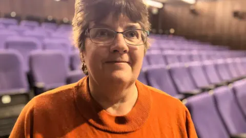 Jon Wright/BBC Joanne Shackle smiles at the camera. There are blue cinema seats behind her. She has brown hair and is wearing glasses and an orange top.