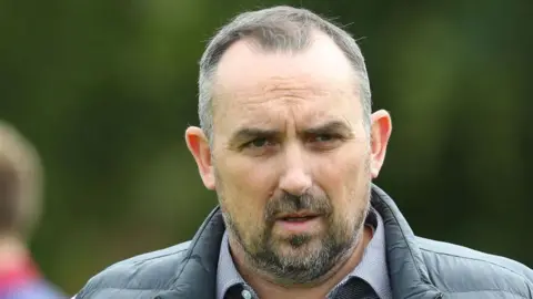 Getty Images Kelvin Thomas wears a black gilet over a blue and white chequered shirt. He has a black and white beard and short greying hair.