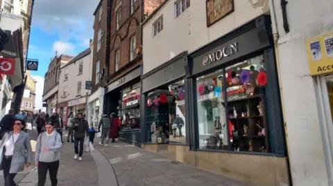 Silver Street which is a pedestrianised road on a hill, busy with people walking up and down in. It is lined with shops on both sides, the one in the foreground has a black front and is called @Moon, but the second o is a crescent moon. It has bunting in the window. There is also a red Vodaphone sign and Body Shop sign on the left side of the street.