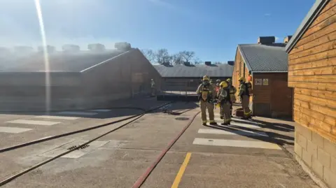 A number of firefighters stand on a courtyard in the middle of a farm site with several buildings around them. The building on the left is emanating white smoke.