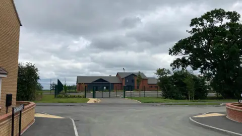 A community centre and car park in front surrounded by green fences and trees. 