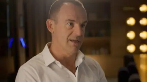 Head and shoulders shot of Martin Lewis looking and talking to side of camera. He has short cropped hair and his wearing a white shirt with the top few buttons open. He is sitting on a chair in what looks like a boardroom, with a long table with chairs on one side and lit light bulbs at the end. 
