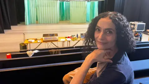 Dominic King/BBC A woman with short dark hair is sitting at a theatre, with the stage behind her. She is looking at the camera with her hand resting under her chin.