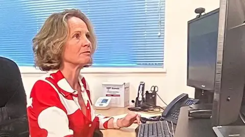 A woman doctor is sat at a computer. She has short blonde hair and is wearing a red and white dress. She is looking at the computer screen.