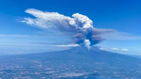 A picture of Mount Etna erupting from a plane. There is grey and black cloud coming out of the top of the volcano. The sky is blue. The ground is green and grey. 