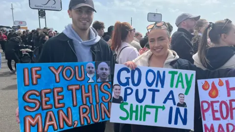 Emma Kruszynski and Jack Penfold, with large crowds visible behind them. Jack Penfold carries a blue sign that says "if you see this man, run" while Emma Kruszynski has a sign that reads "go on Tay, put a shift in".