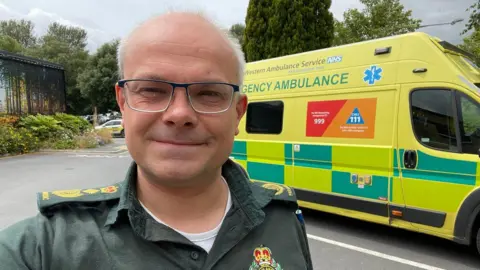 A selfie of Dr John Martin wearing a green paramedic uniform and standing in front of an ambulance, which is hi-vis yellow with green decals down the side.