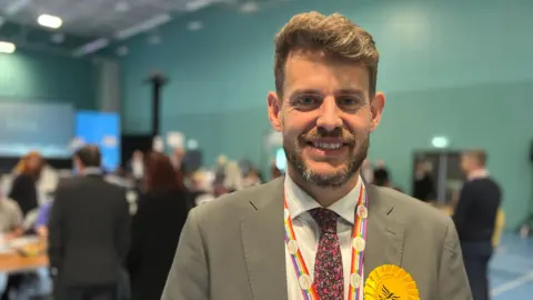 A man in his thirties in a suit and floral-patterned tie smiling at the camera. People are out of focus in the background in a sports hall.