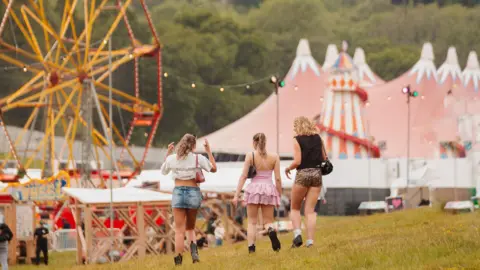 Eljay Three women walk towards fairground rides in the distance, including a ferris wheel and helter skelter.