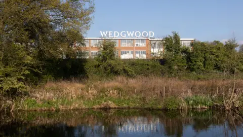 Getty Images The Wedgwood factory building, image taken from across the canal. The Wedgwood name is written in large white lettering above the building and reflected in the canal water. Grass and bushes are at the side of the canal, and a low wall. The two-storey building has large glass windows running along it.