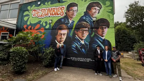 The Rocks Management A mural depicting members of the music group The Zombies on the side of a building with members of the band standing in front of it. The mural is light green and dark green and depicts give men wearing blue velour or velvet jackets. They have 60s style hair and the text is written in a 1960s-style font.