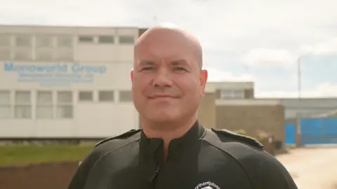 Ant Saddington/BBC Daniel Retter with very short light-coloured hair smiling at the camera while wearing a black shirt with a fire service logo on it. There is a two-storey white building behind him with small windows and Monoworld Group in blue writing.