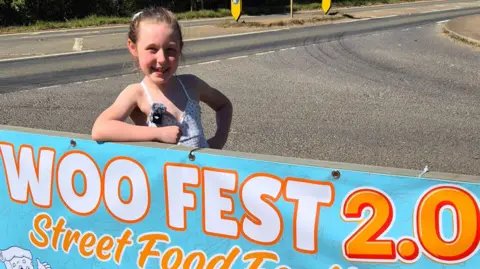 Woo_reviews Woo standing behind a banner-style sign that says WOO FEST 2.0. She is smiling, and has one arm resting in it. A road is behind her. She has her hair tied back and is wearing a white and blue dress.