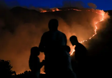 ESA ALEXANDER / REUTERS Silhouettes of people are visible in the foreground. Below them a fires curves along the rim of a mountain.