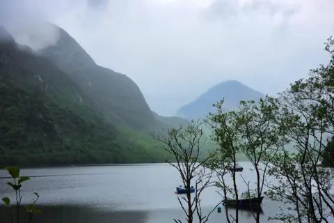 Pathum Vithana A calm loch is beneath dramatic misty green hills. There are three small boats on the water and some trees in the front corner of the scene.