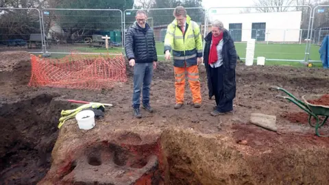 Central Bedfordshire Council Two men, one dressed in black and another wearing high-vis protection on top of his suit, are pointing at the dig site. A woman wearing a long black coat and a red scarf is stood next to them