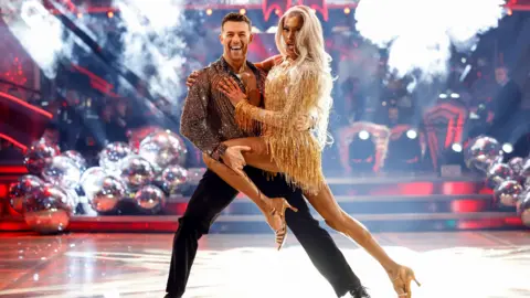 A man and woman dancing while on the Strictly Come Dancing stage. There are red lit steps behind them with giant silver baubles down either side. The man is wearing a sparkly shirt and black trousers and he is holding the woman's left leg across his body. The woman has long light blonde hair and is wearing a short golden dress with tassles. 