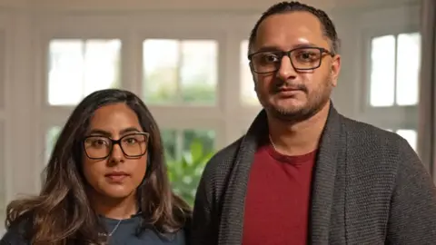 Amarjit and Mandip pose for a photo. They are inside a house. Mandip is wearing a grey cardigan and has glasses while Amarjit also has glasses and brown hair.