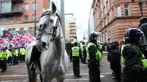 EPA/Shutterstock A police officer is on a police horse which has a protective clear shield over its eyes in a city centre street. Around two other police officers are on horseback behind and multiple police officers are wearing helmets. A crowd of people waving union jack flags can be seen on the left hand side.