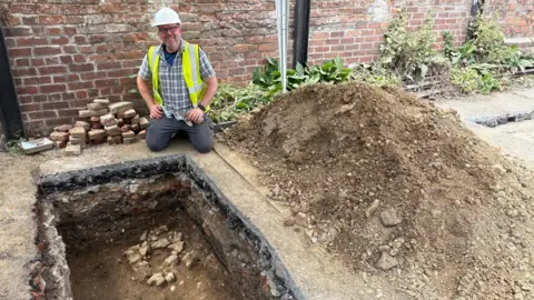 A man wearing a high vis jacket and hard hat is kneeling next to a small pit with a large pile of dirt nearby