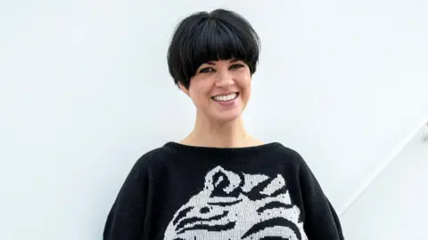Dawn O'Porter stood in front of a white background. She has a black and white jumper on and a short black bob hairstyle. 