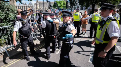A number of police officers on a road with some members of the public taking photos and filming. The police appear to be carrying a person although only their foot can be seen in the image.