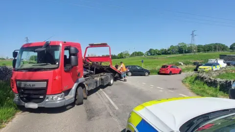 Derbyshire Police Red vehicle on the left of the image with a police vehicle on the right. There are green fields in the distance and blue sky above