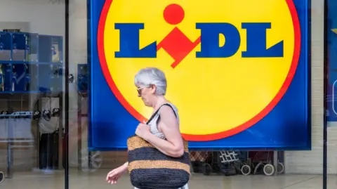 A woman with short grey hair wearing a grey sleeveless top carrying a black and brown wicker bag. She is walking past a large LIDL sign on a window.