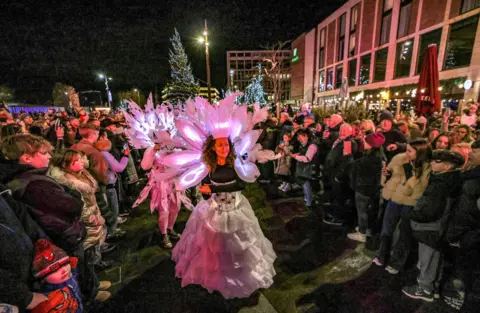 Sunderland City Council Crowds look on as entertainers make their way to Keel Square as part of the parade. A woman is wearing an illuminated white headdress and flowing white skirt.