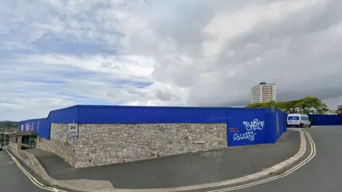 Google A Google street view screen grab of the derelict hotel site, hidden by tall navy panels, cloudy skies