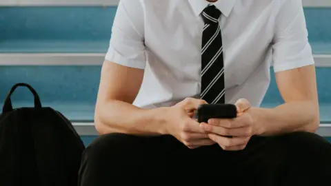 A stock image of a boy sitting on blue stairs. He is wearing a white short-sleeved shirt, with a black and white tie and black trousers. He is holding a phone in both hands. A black rucksack is sitting on the stairs on the left of the image.