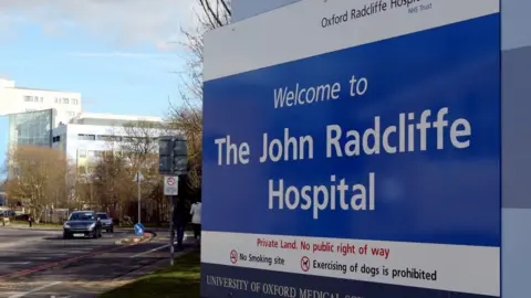 File photo of the entrance to the John Radcliffe Hospital in Oxford. It is in large white writing on a blue NHS sign, with a large blue building behind the sign.