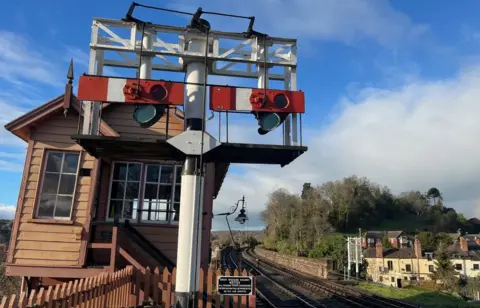 A light brown signal box is on the left, along with signals. Track is near the middle of the photo, with houses and trees to the right.