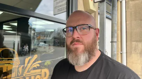 A man with a shaved head and a beard. He is wearing glasses.